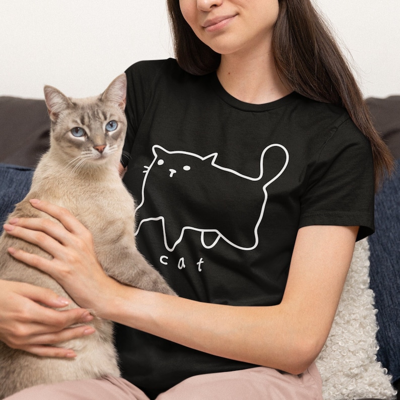 May include: A woman wearing a black t-shirt with a white outline of a cat and the word "cat" printed on it. She is holding a light brown cat in her arms.