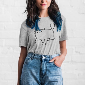 May include: A gray t-shirt with a black line drawing of a cat and the word "cat" printed on the front. The model is wearing blue jeans.