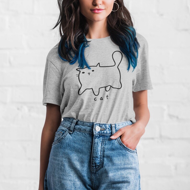 May include: A gray t-shirt with a black line drawing of a cat and the word "cat" printed on the front. The model is wearing blue jeans.