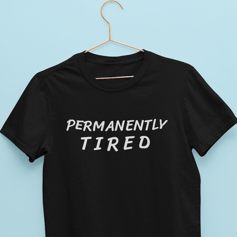 May include: Black t-shirt with white text that reads "PERMANENTLY TIRED" on a hanger against a light blue background.