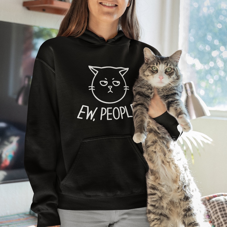 Pu&ograve; includere: Felpa con cappuccio nera con un viso di gatto bianco e il testo "Ew, People" stampato sul davanti. Una persona indossa la felpa e tiene un gatto tigrato.