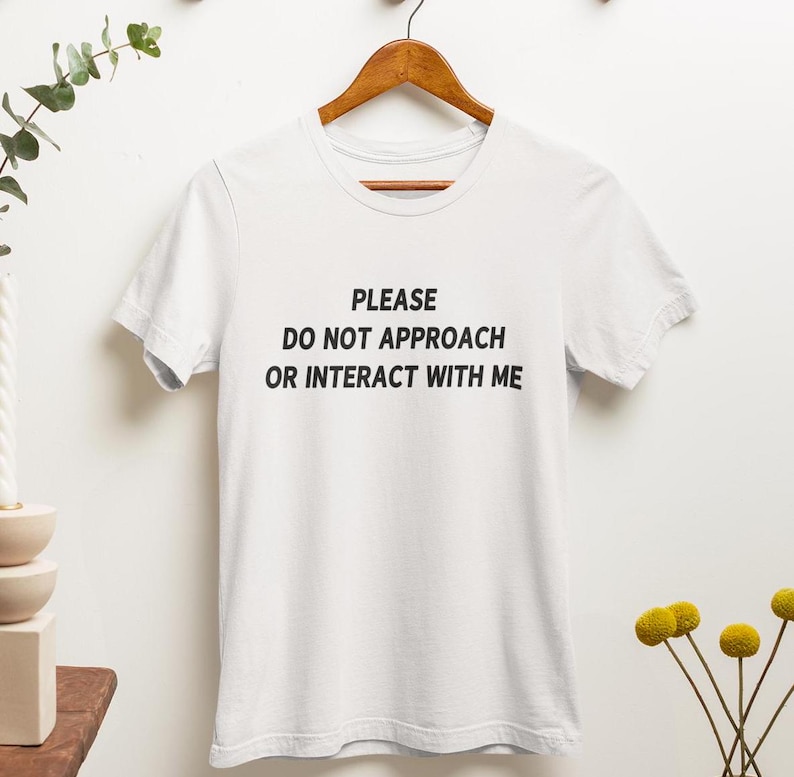 K&ouml;nnte beinhalten: Wei&szlig;es T-Shirt mit dem Text "PLEASE DO NOT APPROACH OR INTERACT WITH ME" in Schwarz gedruckt.