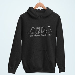 Op de afbeelding: Een donkergrijs hoodie met een witte afbeelding van vier katten en de tekst "un deux trois cat".