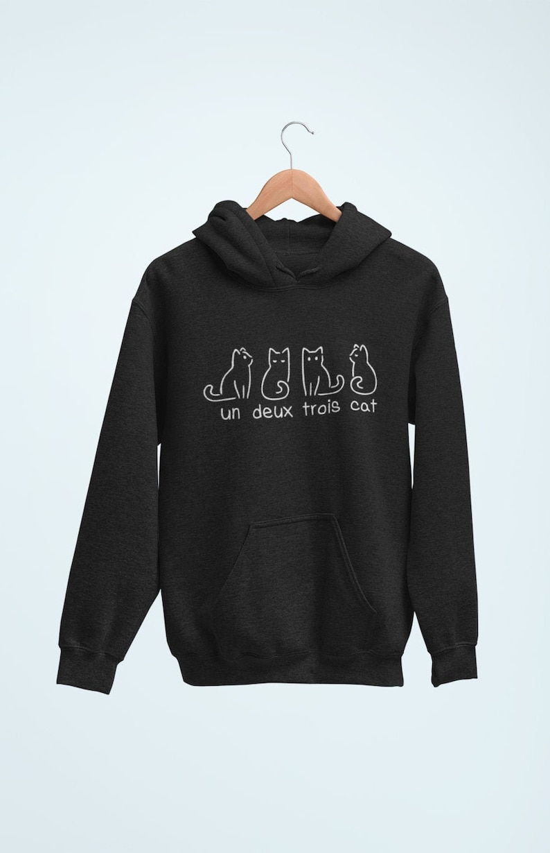 Op de afbeelding: Een donkergrijs hoodie met een witte afbeelding van vier katten en de tekst "un deux trois cat".