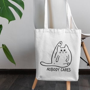 May include: White tote bag featuring a black line drawing of a cat with its leg raised in the air. The text "NOBODY CARES" is printed below the cat.