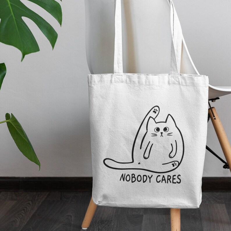 May include: White tote bag featuring a black line drawing of a cat with its leg raised in the air. The text "NOBODY CARES" is printed below the cat.