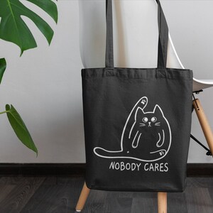 May include: Black tote bag with a white outline of a cat and the text "Nobody Cares" printed on the front.