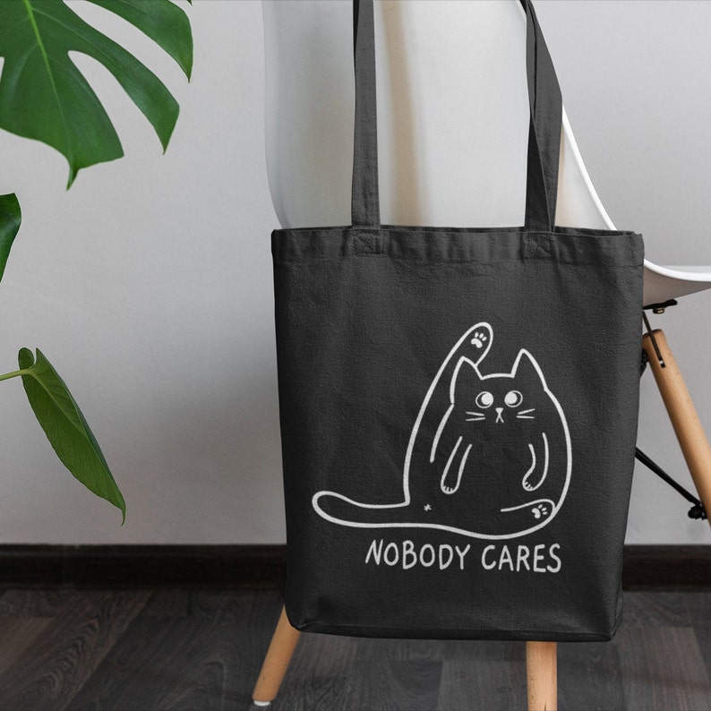 May include: Black tote bag with a white outline of a cat and the text "Nobody Cares" printed on the front.