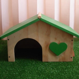 Può includere: Una casetta di legno per animali domestici con un tetto verde e un cuore verde ritagliato sul davanti. La casetta è su una superficie di erba artificiale verde.