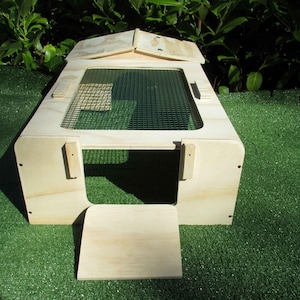 Può includere: Un habitat in legno per piccoli animali con un top in rete metallica e una porta a cerniera. La struttura è realizzata in legno di colore chiaro e poggia su una superficie verde. L'habitat ha una forma rettangolare con tetto spiovente.