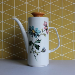 May include: A white ceramic coffee pot with a floral design featuring blue, pink, and green flowers. The pot has an orange lid and a spout on the side.