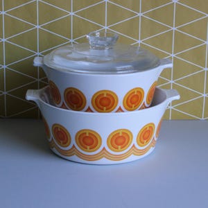May include: Two white vintage casserole dishes with orange and yellow circular designs. One dish is stacked on top of the other. The top dish has a clear glass lid with a small handle. The dishes have side handles.