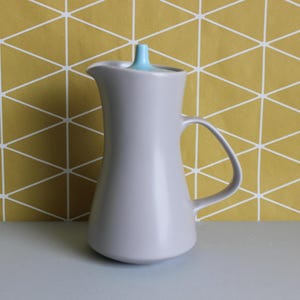 May include: A grey ceramic jug with a curved handle and a light blue lid. The jug has a slender, elegant shape and a spout for pouring. The background features a yellow and white geometric pattern.