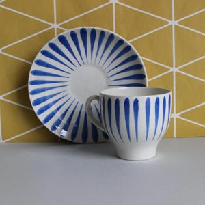 May include: White ceramic teacup and saucer set with a blue stripe design. The cup has a handle and a rounded shape. The saucer is round. The background is a yellow geometric pattern. The cup and saucer are suitable for tea or coffee.