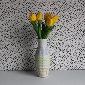 May include: A ceramic vase with a woven design in pastel colours holds a bouquet of yellow tulips. The vase has a white top and is decorated with horizontal bands of lavender, green, and yellow. The tulips have green stems and leaves.
