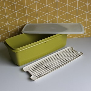 May include: A retro green rectangular food container with a white grid insert and a clear lid. The container is open, revealing the inside. The background features a yellow and white geometric pattern.