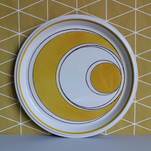 May include: A white ceramic plate with a yellow and brown geometric design. The design features concentric circles and a crescent shape.