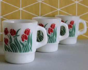 May include: Three white ceramic mugs with a red and green tulip design. The mugs are stacked on top of each other.