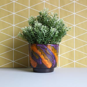 May include: A ceramic plant pot with a striking design. The pot has a black base and a body with streaks of orange, purple, and yellow. It contains a small, green plant with white accents. The background is a gold geometric pattern, adding to the visual appeal.