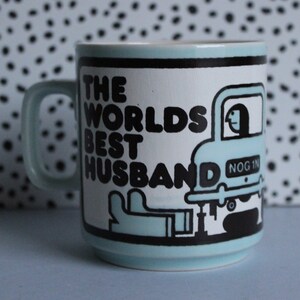 May include: A light blue ceramic mug with a black and white graphic of a car with the text "The Worlds Best Husband" and "Nog 1" on the side.