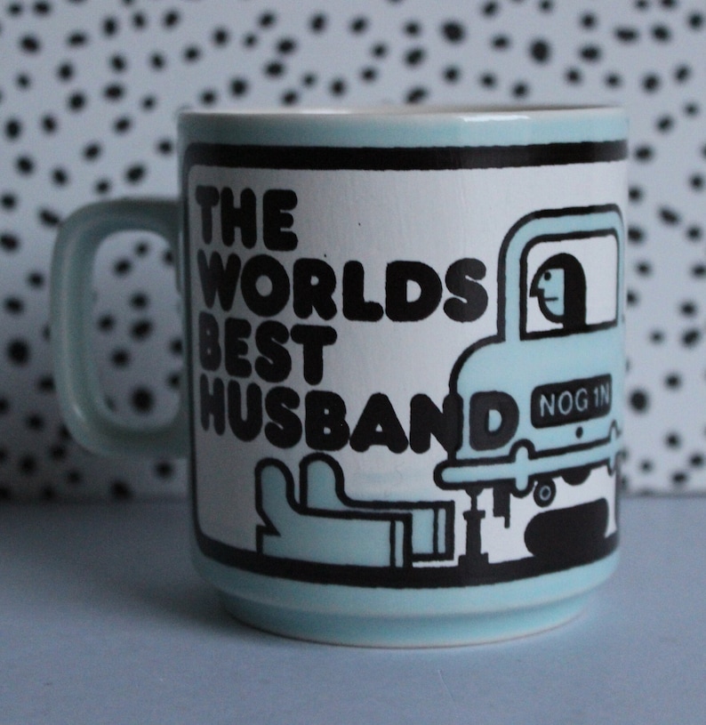 May include: A light blue ceramic mug with a black and white graphic of a car with the text "The Worlds Best Husband" and "Nog 1" on the side.