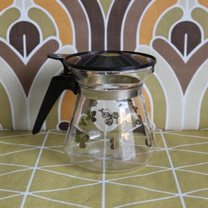May include: A vintage glass coffee pot with a black handle and lid. The clear glass is decorated with gold and green butterfly and clover designs. The coffee pot has a silver band around the middle.