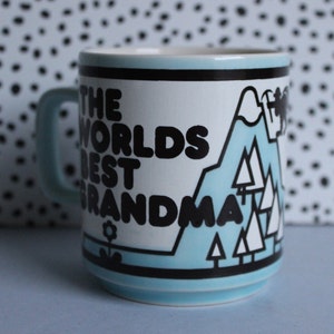 May include: A blue and white ceramic mug with black text that reads "The Worlds Best Grandma" and a graphic of a mountain range with trees.