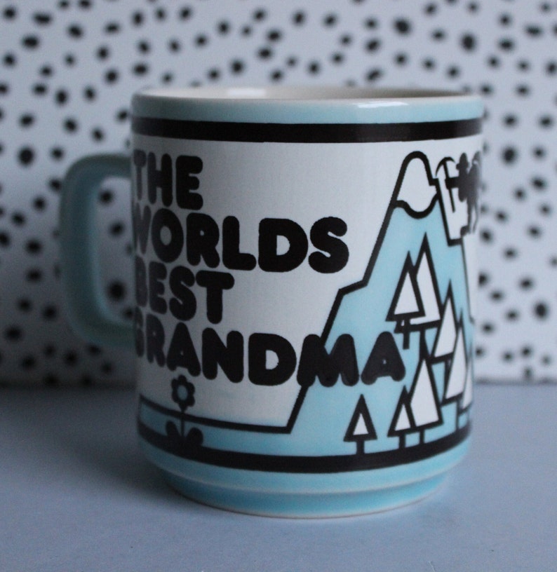 May include: A blue and white ceramic mug with black text that reads "The Worlds Best Grandma" and a graphic of a mountain range with trees.