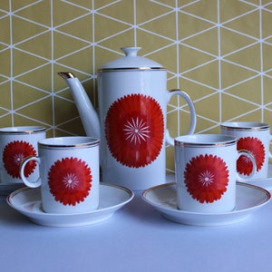 May include: A white ceramic coffee pot and four matching cups with red floral designs and gold trim. The set is on a light blue surface with a yellow geometric pattern background.