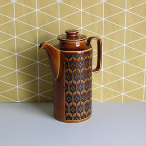 May include: A tall, cylindrical ceramic coffee pot featuring a glossy brown glaze on the lid, handle, and spout. The main body displays a dark brown and orange geometric pattern consisting of repeating vertical links and small star-like shapes.