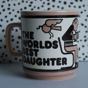 May include: A pink ceramic mug with a black and white illustration of a person sitting on a chair and a bird flying. The mug has the text "The Worlds Best Daughter" in black letters.