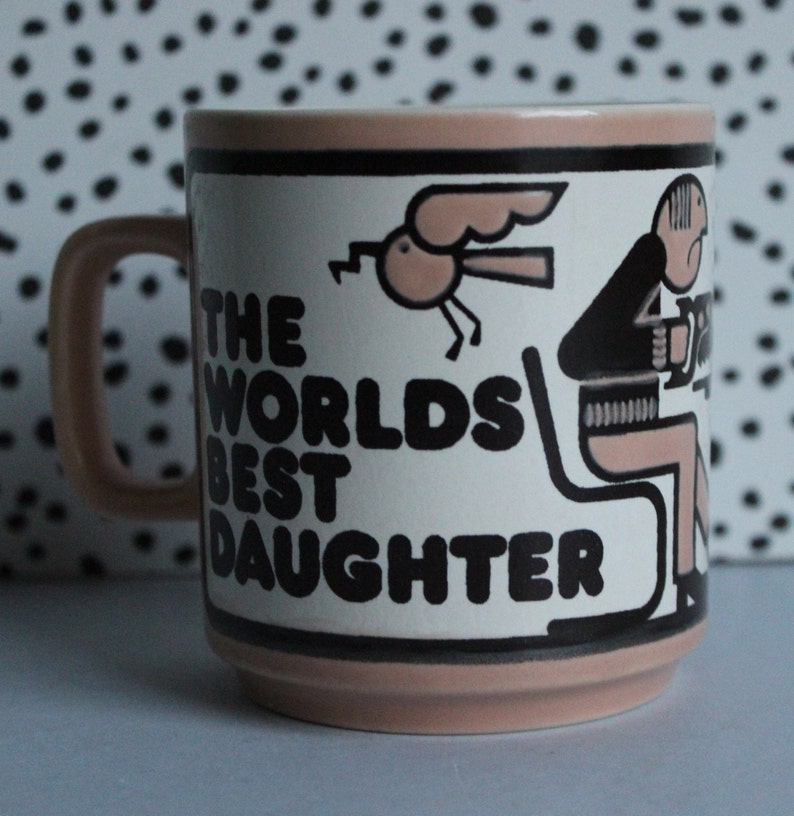 May include: A pink ceramic mug with a black and white illustration of a person sitting on a chair and a bird flying. The mug has the text "The Worlds Best Daughter" in black letters.