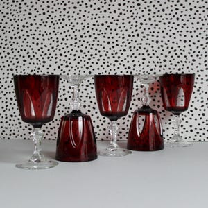 May include: Set of five red wine glasses with a clear stem and base. The glasses have a cut glass design and are arranged in a row on a white surface.