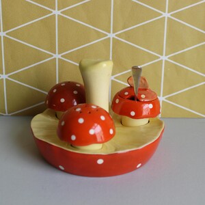 May include: A novelty mushroom-shaped condiment set. The set includes a salt and pepper shaker and a sugar bowl, all with red caps and white spots. The set is on a yellow base. The sugar bowl has a small spoon.