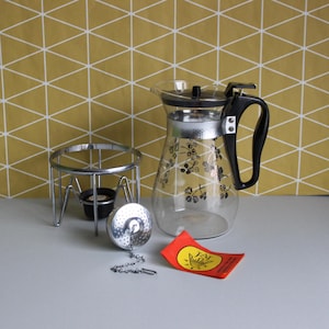 May include: A vintage glass teapot with a black handle and floral design, alongside a metal tea warmer, a tea infuser, and a red tag with yellow text. The teapot has a silver rim and a black lid.