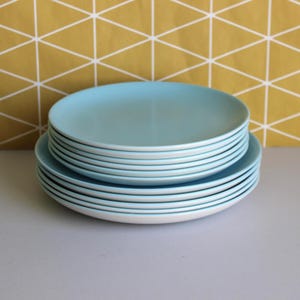 May include: A stack of round ceramic plates featuring a smooth, light blue top surface and a crisp white underside. The plates are arranged in two distinct tiers, with a smaller stack of light blue plates resting on top of a larger stack of plates.
