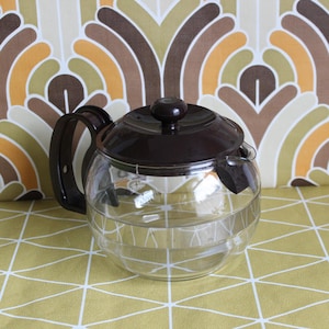 May include: A clear glass teapot with a dark brown lid, handle, and spout. The teapot has a rounded shape and a white geometric pattern. The background features a retro-style patterned fabric in shades of brown, orange, and beige.
