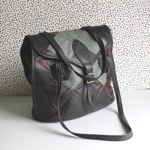 May include: A patchwork leather shoulder bag in black, grey, and burgundy. The bag has a flap closure with a buckle and a long adjustable strap. The design is a mix of geometric shapes.