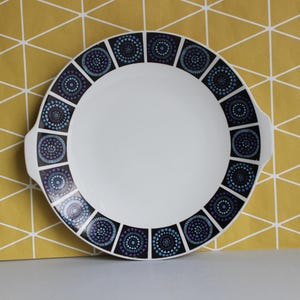 May include: White ceramic serving platter with a geometric border design in shades of blue, purple, and black. The platter has two small handles and a large, empty centre. The design features circular patterns within square frames.