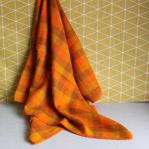 May include: A fabric textile featuring a plaid pattern in shades of bright orange, golden yellow, and deep brown. The material is draped to show its soft texture and woven construction, displaying a consistent grid of intersecting coloured lines.