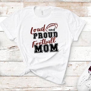 May include: White t-shirt with a red and black graphic that says "Loud and Proud Football Mom" with a red football graphic.