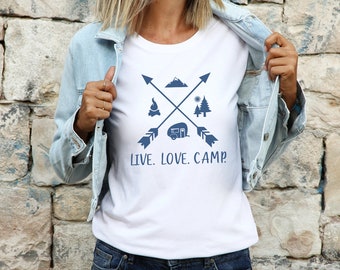 May include: White t-shirt with a blue graphic design featuring crossed arrows, a mountain, a campfire, a camper, and two pine trees. The text "LIVE. LOVE. CAMP." is printed below the graphic.