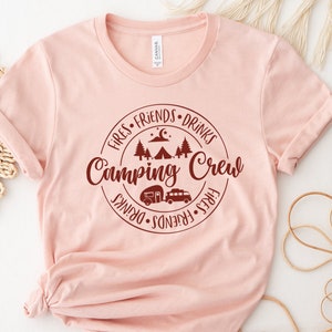 K&ouml;nnte beinhalten: Ein pinkfarbenes T-Shirt mit einem roten Kreis-Motiv, auf dem "Fires, Friends, Drinks, Camping Crew, Fires, Friends, Drinks" steht. Das Motiv enth&auml;lt auch ein Zelt, B&auml;ume, einen Mond und einen Wohnwagen.