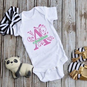 May include: A white baby onesie featuring a pink letter A and green floral design. The text "Anastasia" is written below the letter. The onesie is displayed on a wooden background with a black and white headband, a panda toy, and gold and black striped baby shoes.