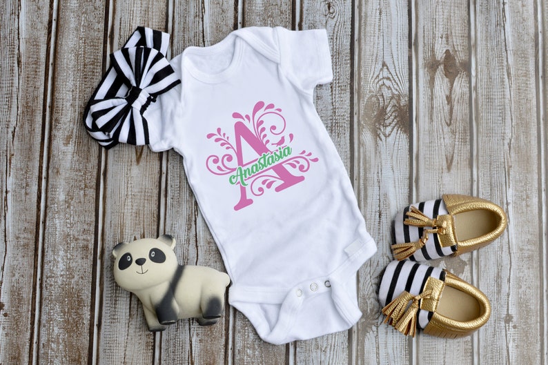 May include: A white baby onesie featuring a pink letter A and green floral design. The text "Anastasia" is written below the letter. The onesie is displayed on a wooden background with a black and white headband, a panda toy, and gold and black striped baby shoes.