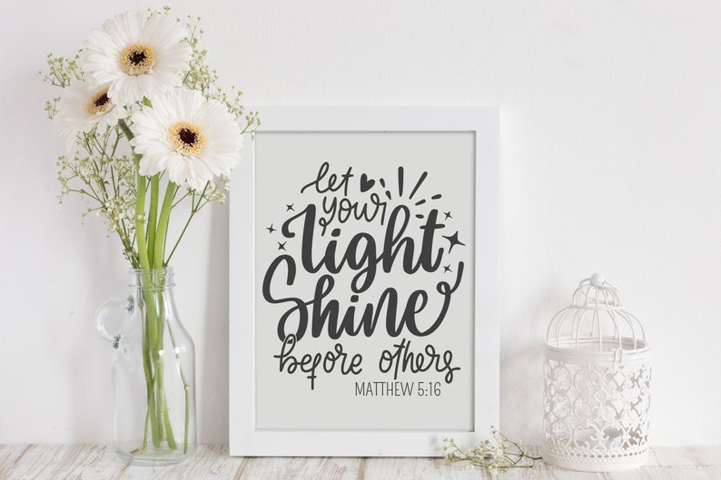 May include: A white framed print with the words "let your light shine before others" in black script. The image is displayed with white gerbera daisies in a clear glass bottle and a decorative white birdcage.