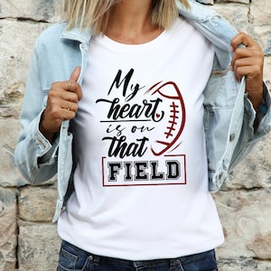 May include: A white short-sleeved t-shirt featuring a graphic design with a football illustration and the black and maroon text "My heart is on that FIELD". The shirt is worn under a light blue denim jacket, showcasing a casual layered look.
