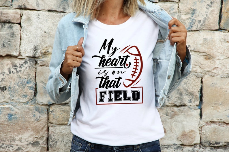 May include: A white short-sleeved t-shirt featuring a graphic design with a football illustration and the black and maroon text "My heart is on that FIELD". The shirt is worn under a light blue denim jacket, showcasing a casual layered look.