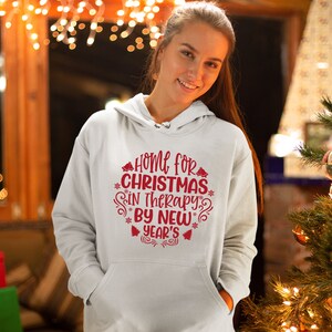 May include: White hoodie with red text that reads "Home for Christmas in Therapy by New Year's". The text is surrounded by snowflakes and bells.
