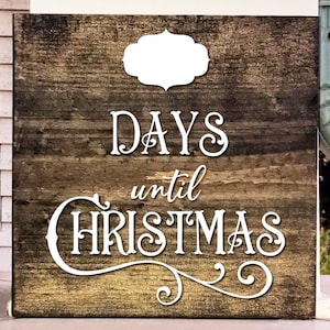 Può includere: Un cartello di legno con una finitura marrone invecchiata. Il cartello ha testo bianco che recita "DAYS until CHRISTMAS" con un fiocco decorativo. C'è un'etichetta bianca vuota nella parte superiore del cartello.